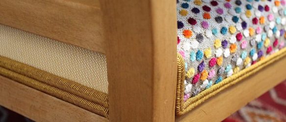 Zoom sur un montant du petit voltaire enfant. on voit le bois clair, un bout du dos avec un tissu qui rappel le bois, un bout de l'assise en tissu Casal à pois multicolore sur fond écru ainsi que le double passepoil tressé de fnition de couleur jaune/doré